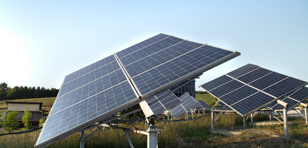 Ground-mounted solar panels in a field, tilted at different angles under a clear sky, with trees and a building in the background.