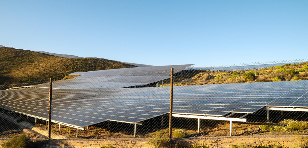 Solar panels on a hillside behind a fence under a clear sky, stretching into the distance.