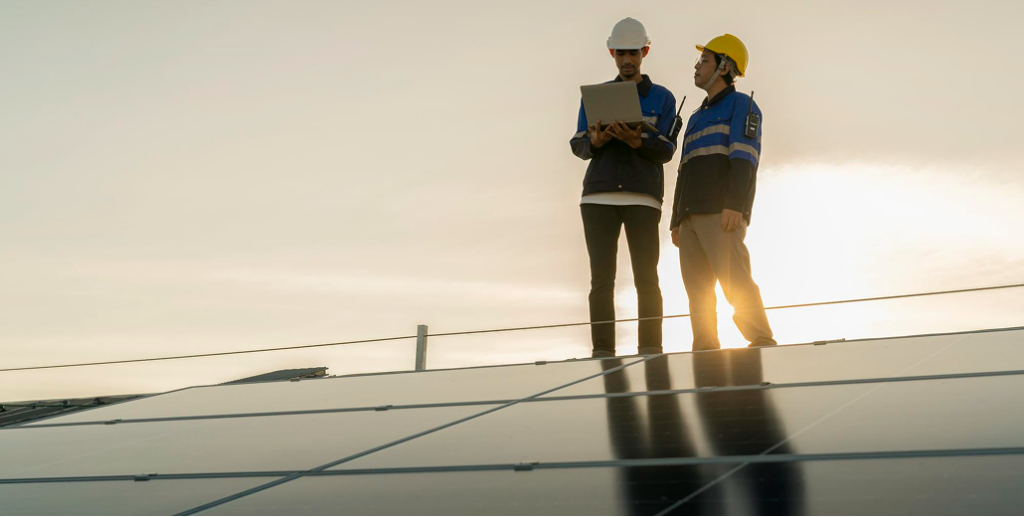 Two construction workers in hard hats and workwear on a solar panel array, one using a laptop.