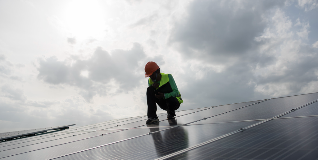 An installer wearing an orange hard hat and high-vis vest inspects a large array of solar panels under a cloudy sky.