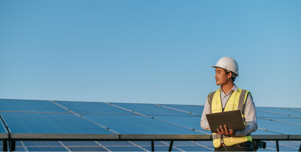 A solar engineer in a hard hat and safety vest holds a laptop in front of a ground-mounted solar panel array.