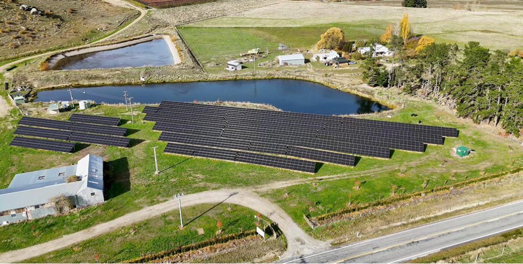 Aerial view of a large ground-mounted solar panel array on a green hillside next to a blue pond on a farm.
