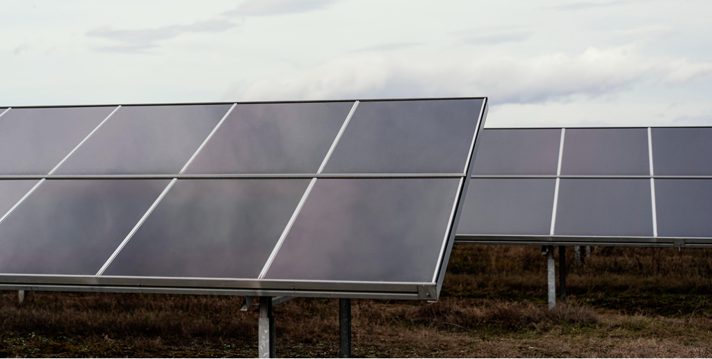 Ground-mounted solar panels angled on a metal frame over dry, brown grass under a cloudy sky.