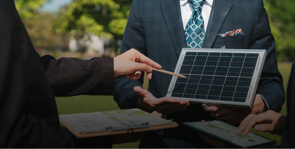 Business professionals in suits holding and pointing to a small demonstration solar panel during a meeting.