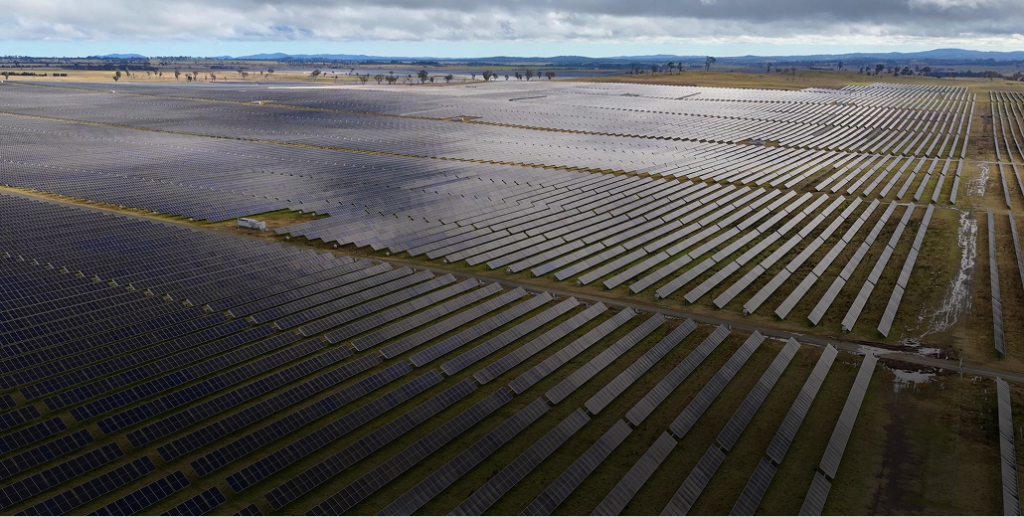 Aerial view of massive rows of dark solar panels stretching across rolling hills under a cloudy sky.