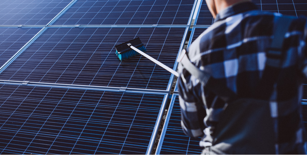 A worker in a plaid shirt cleans a large array of blue solar panels using a long-handled brush.
