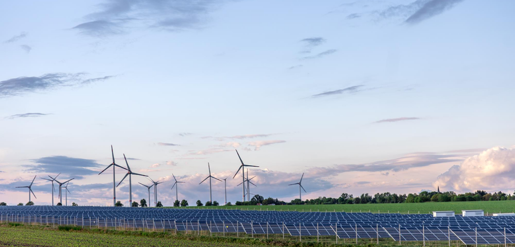 A wide field of ground-mounted solar panels stretching out in front of several large wind turbines under a cloudy sky.